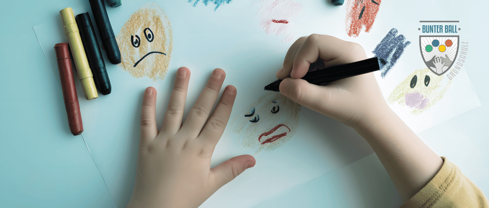 Ein Blatt Papier ist zu sehen, auf dem Wachsmalstifte liegen und eine Kinderhand Gesichter mit unterschiedlichen Gefühle darauf zeichnet