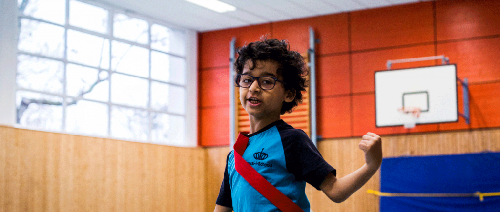 Ein Kind im blauen T-Shirt läuft durch eine Sporthalle. Das Kind hat braune Locken und eine Brille.